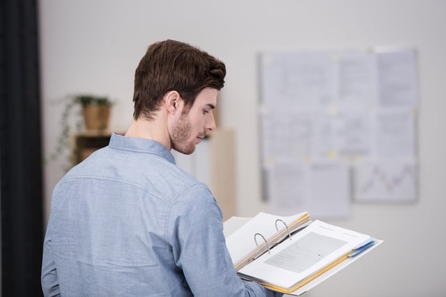 Young man standing doing research in the office reading from a hardcover book balanced on his palm, view over his shoulder from the rear.jpeg